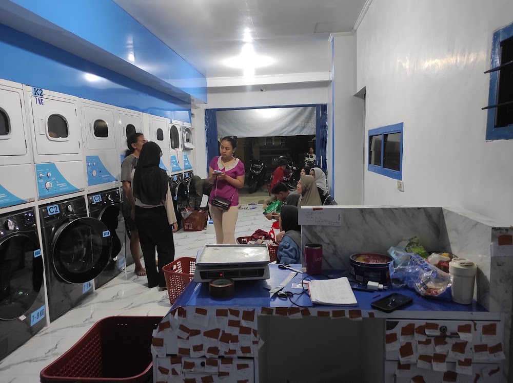 Customers and staff waiting inside a busy laundromat near a service desk and rows of laundry machines.