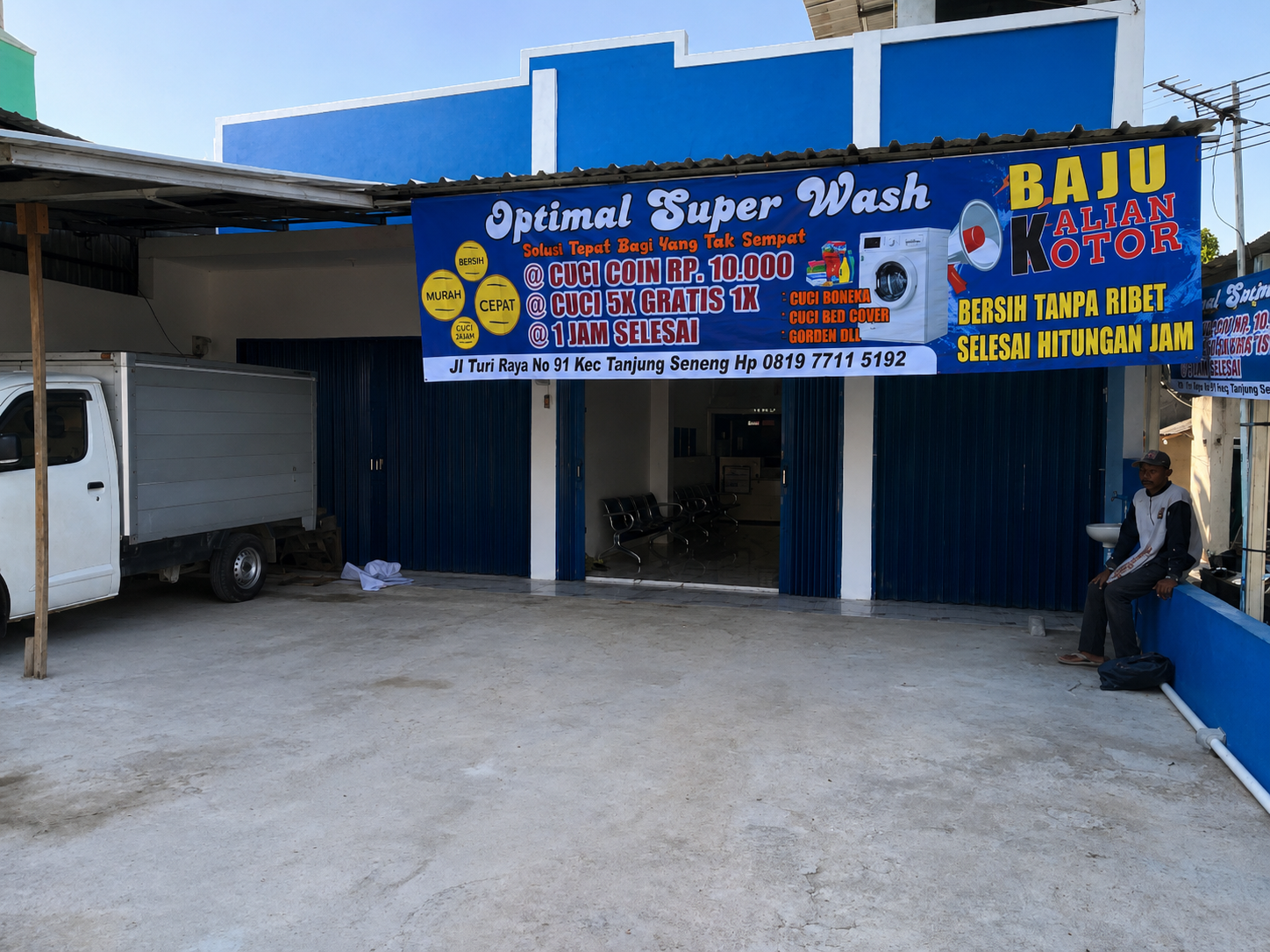The exterior storefront of Optimal Super Wash featuring a large promotional banner, an open entrance, a small delivery truck, and a concrete forecourt.